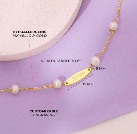 Product shot on a lavender background of the 14 k yellow-gold pearl bracelet with name plate, overlaid with annotations: ‘Hypoallergenic 14 k Yellow Gold,’ ‘5"–Adjustable to 6",’ ‘Pearl: 5 mm,’ ‘Name Plate: 15 mm,’ and ‘Customizable Engraving’ (shown with the name “Anne”).
