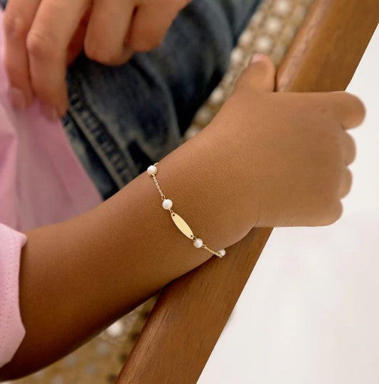 Close-up of a child’s wrist resting on a wooden armrest, wearing the 14 k yellow-gold pearl bracelet with four small white pearls and a central name plate on a delicate chain.