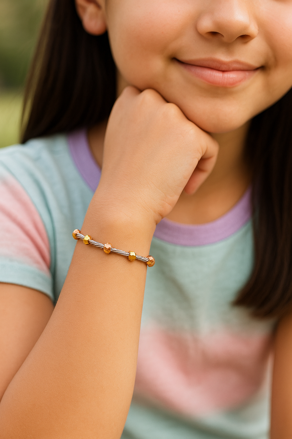Close-up of a tween girl’s left wrist wearing a tri-colour beaded bracelet, featuring twisted silver rods and faceted gold and rose-gold beads, shown against a softly blurred outdoor background