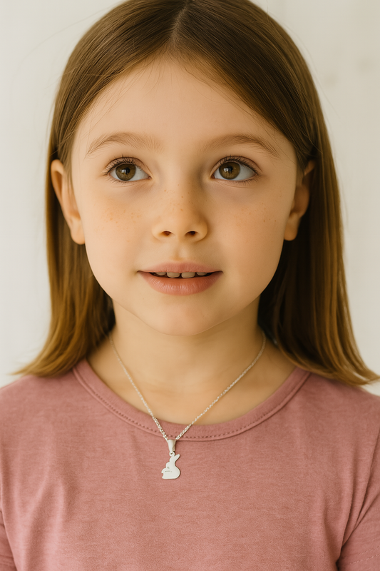 Portrait of a young girl wearing a silver necklace with a pendant shaped like two cuddling rabbits. She has straight brown hair, warm hazel eyes, and a soft smile, dressed in a dusty pink top with a light, neutral background
