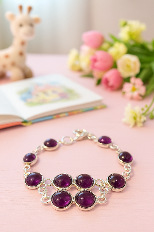 A children’s silver bracelet with seven round purple amethyst cabochons laid out on a pastel pink wooden tabletop; behind it, an open colorful storybook lies blurred to the left, a soft plush giraffe toy sits in the background, and a small bouquet of pink and white flowers rests to the right, creating a warm, playful scene