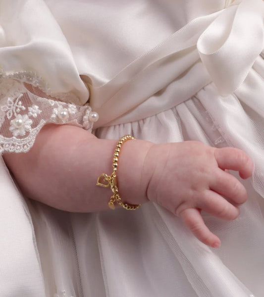 Close-up of a baby’s forearm wearing a delicate gold beaded bracelet with a dangling open-heart charm and small oval tag, contrasted against the baby’s white lace-trim dress sleeve.
