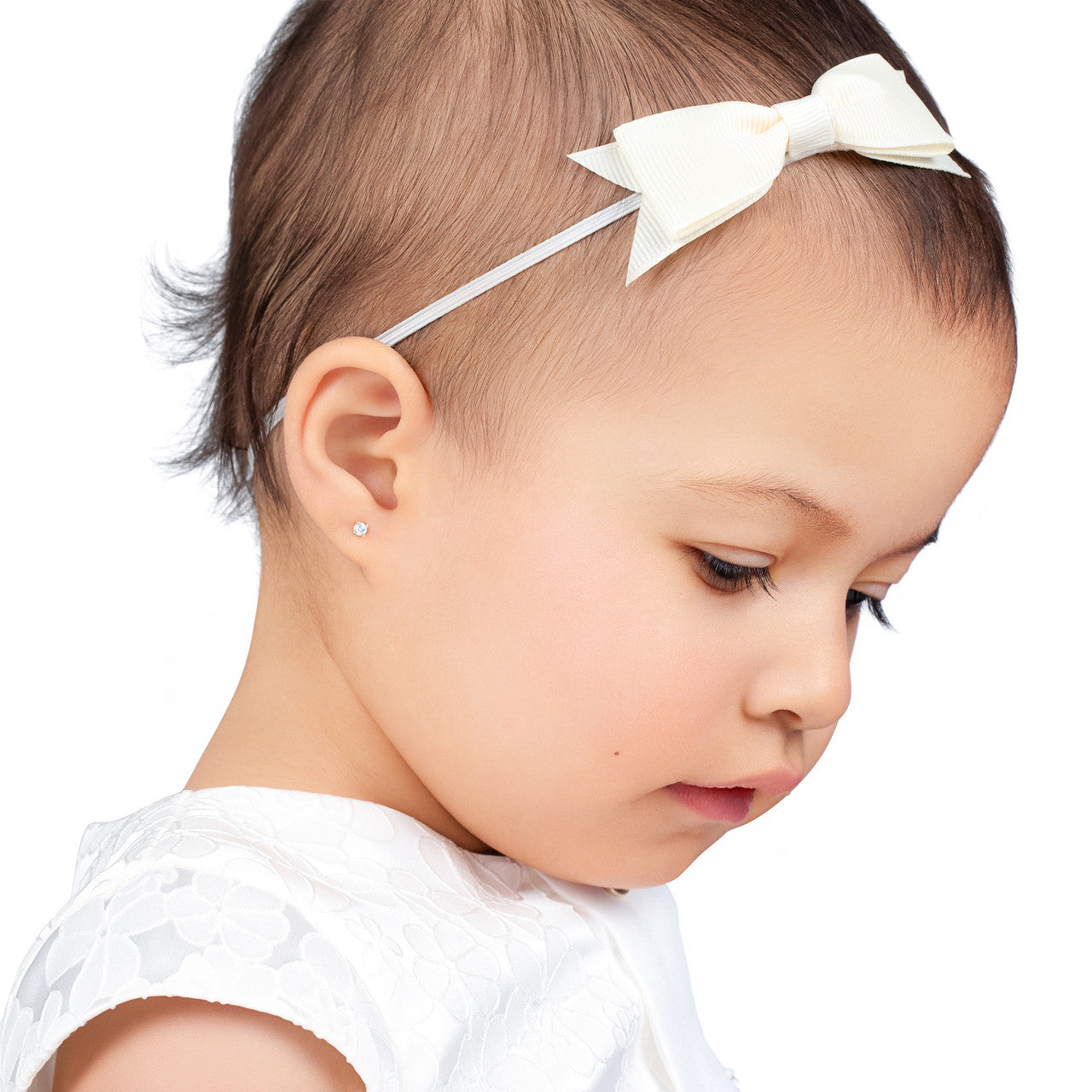 Side profile of a toddler wearing a tiny diamond stud earring, a white floral dress, and a cream bow headband, photographed against a clean white background.