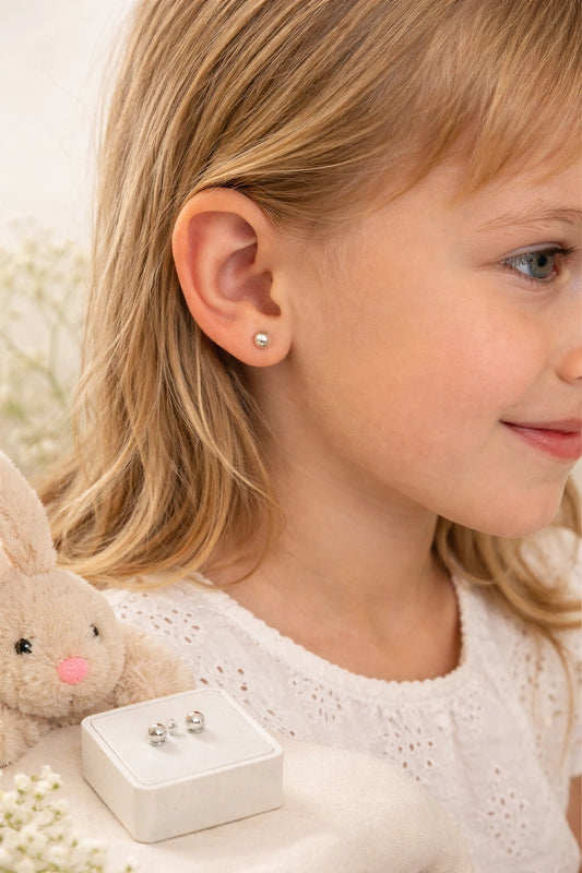 A young girl wearing children's earrings featuring 14k white gold ball studs, with a bunny plush and a box in the background.