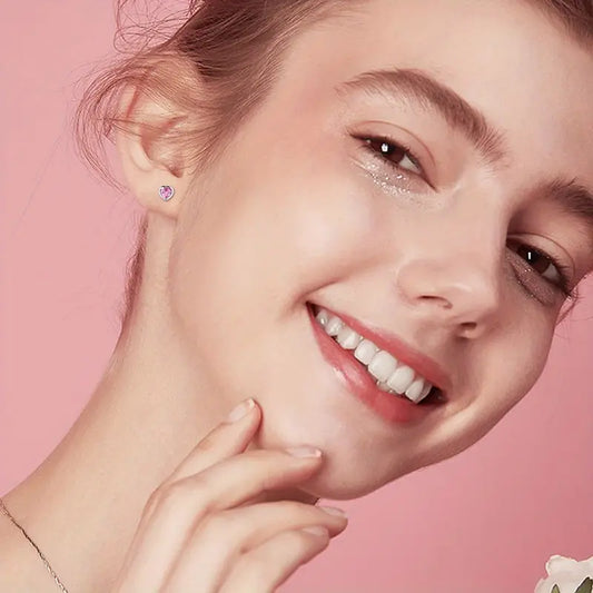Close-up of a smiling young teen girl against a pink background, showcasing a small pink heart-shaped stud earring in her ear and wearing subtle sparkly eye makeup.