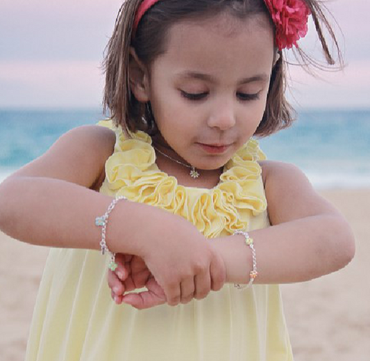 Portrait of a young girl on a sandy beach at sunset, wearing a yellow ruffled dress and a red flower headband, looking down to admire two delicate sterling-silver chain bracelets adorned with enamel daisy charms.