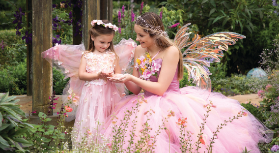 Two girls dressed as fairies in a lush garden. The older girl wears a voluminous pink tulle gown, iridescent butterfly wings, and a delicate floral crown, smiling as she holds out flower petals. Beside her, a younger girl in a matching pink dress with sheer fairy wings and a blossom headband looks down at the petals in her hands, surrounded by blooming flowers and greenery.