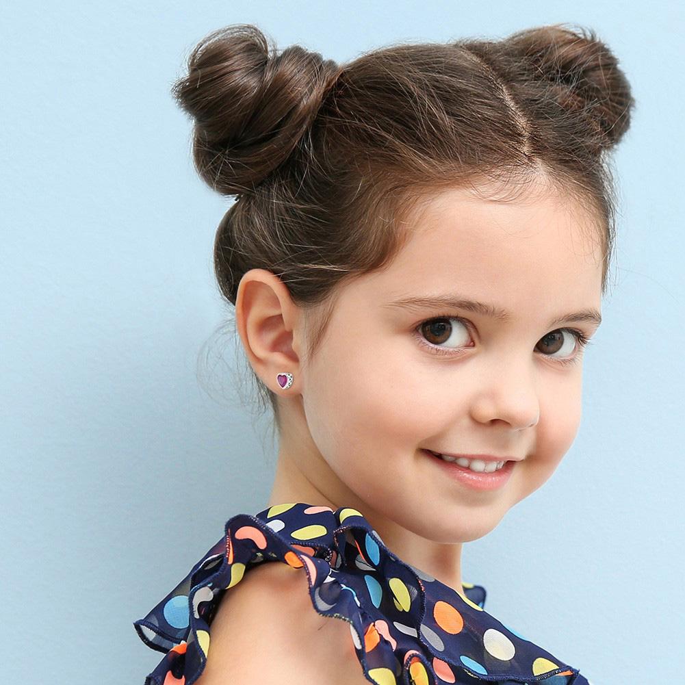 A young girl with brown hair tied up in two knots in a navy frilled top with coloured circles wearing silver heart shaped stud earrings