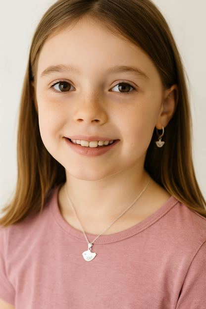 Portrait of a smiling young girl wearing a silver baby bird pendant necklace on a fine chain, styled with a pink t-shirt and softly lit background for a playful, youthful look