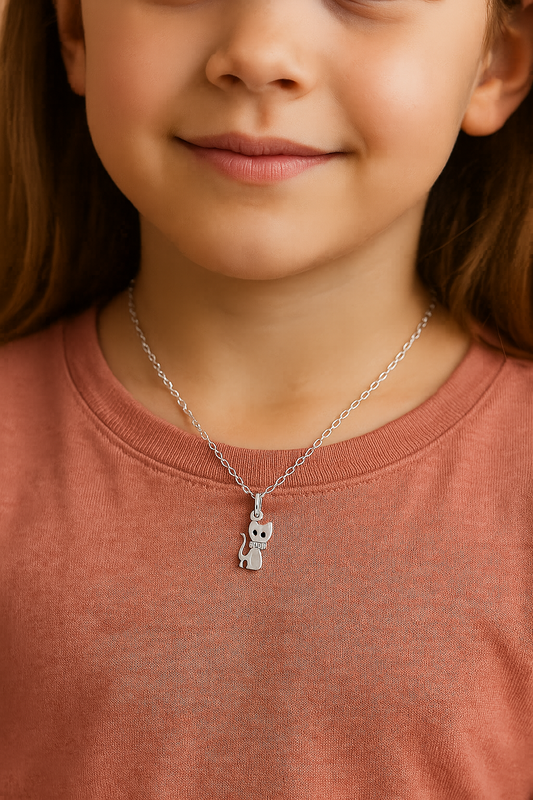 Close-up portrait of a young girl wearing a silver cat pendant necklace with plain black indented eyes. The necklace hangs from a delicate chain and is centred on her neck. She is dressed in a salmon-coloured T-shirt, and the soft lighting highlights her natural smile and warm skin tone with a neutral background.