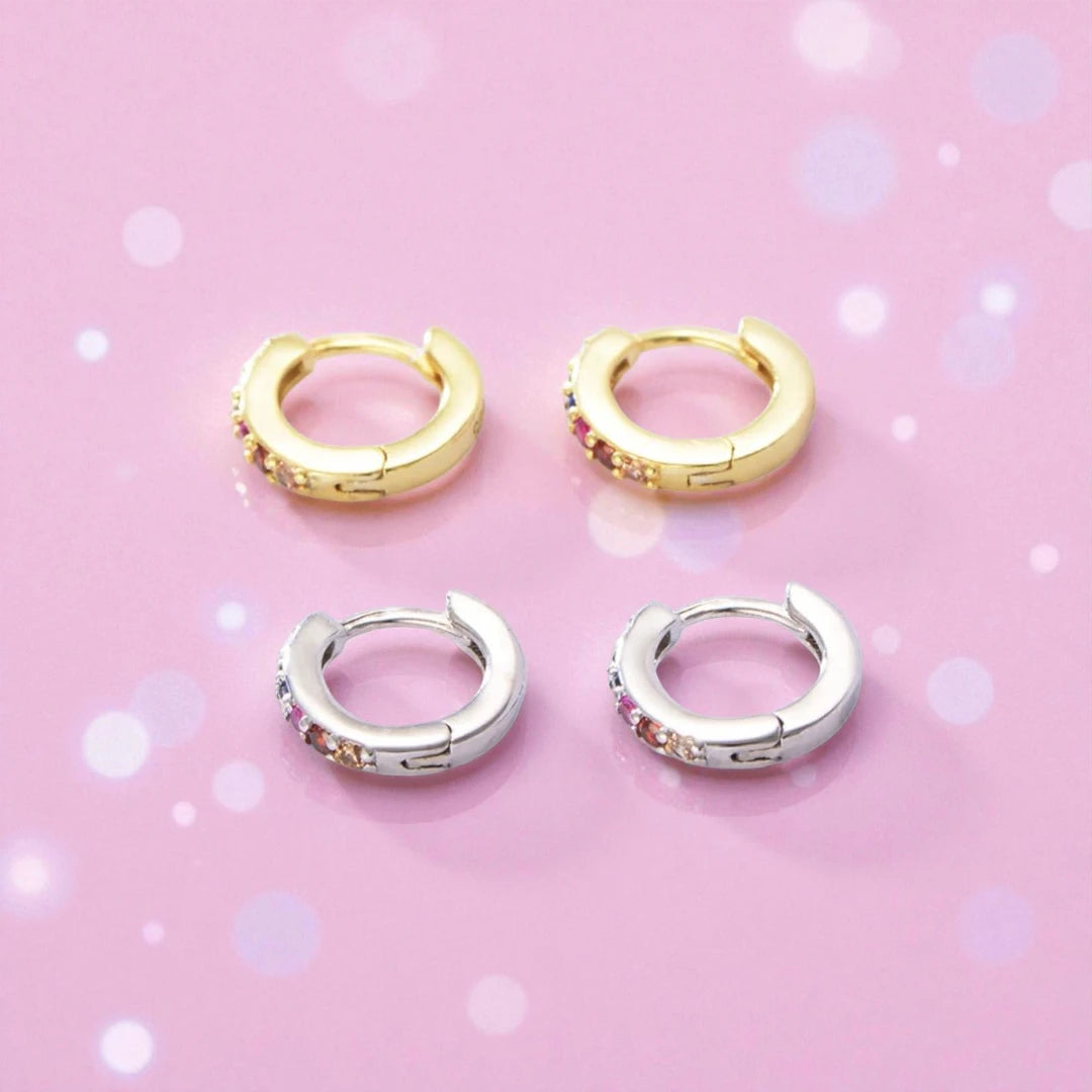Baby hoop earrings in gold and silver with rainbow CZ stones on a pink background.