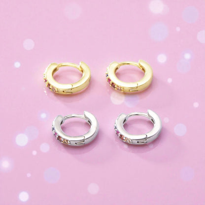 Baby hoop earrings in gold and silver with rainbow CZ stones on a pink background.