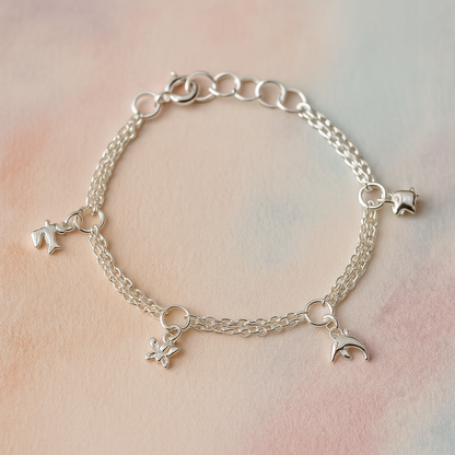 Sterling silver children’s charm bracelet with double chains and four small animal charms—a hummingbird, butterfly, dolphin, and elephant—laid out on a soft pastel watercolour background.