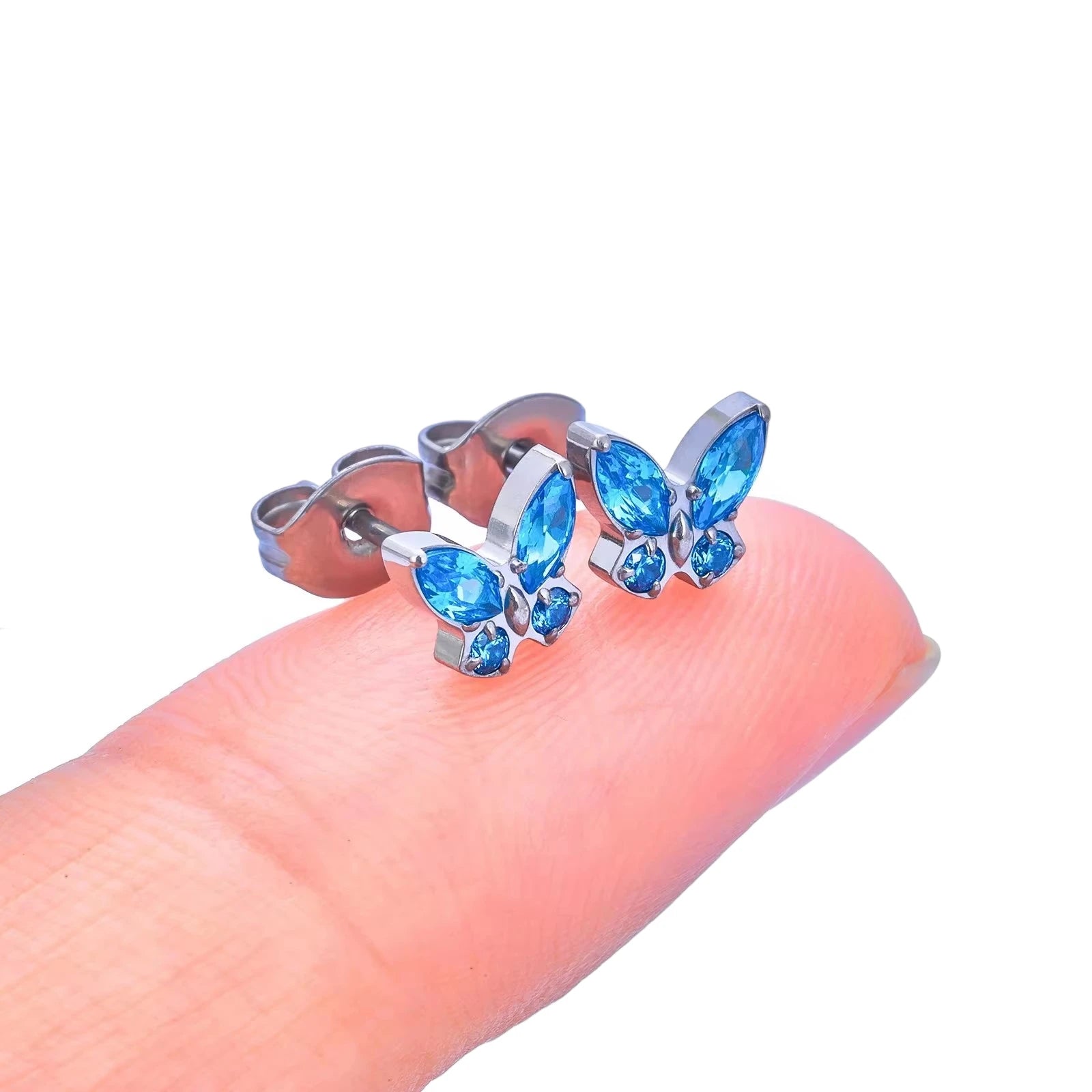 Children's earrings featuring blue CZ crystal butterfly studs on a fingertip, crafted from G23 titanium.
