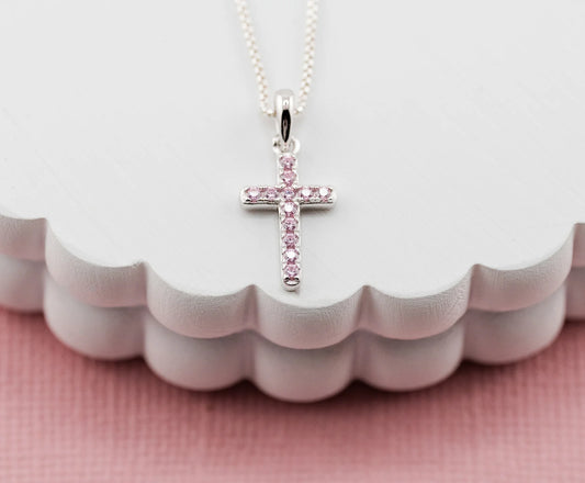 Silver cross necklace with pink stones on a white stand with a pink background