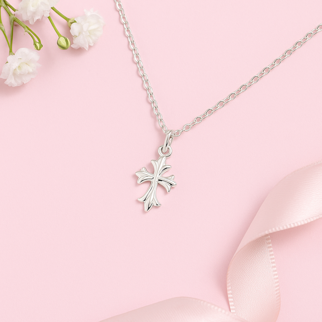 Children's sterling silver fleur de lis cross necklace on a light pink background with flowers and ribbon.