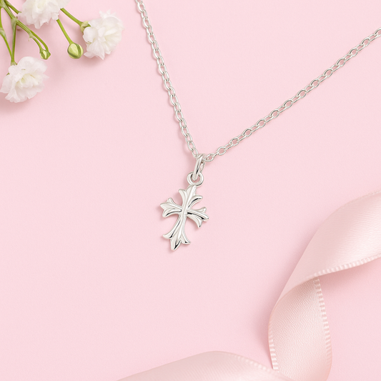 Children's sterling silver fleur de lis cross necklace on a light pink background with flowers and ribbon.