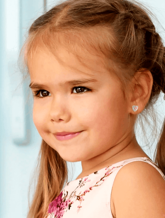 Close-up of a smiling young girl with light brown hair in pigtails, wearing a sleeveless floral pink dress and small silver screw-back heart stud earrings with pink stones, set against a softly blurred pastel background