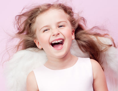 A laughing young girl with long, windblown hair wearing a pale pink sleeveless top and white angel wings, against a pastel pink background, showing small silver stud earrings on her ears.