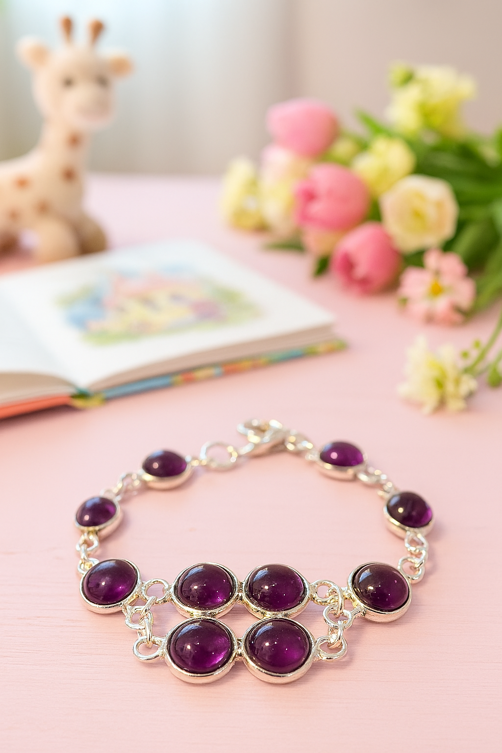 A children’s silver bracelet with seven round purple amethyst cabochons laid out on a pastel pink wooden tabletop; behind it, an open colorful storybook lies blurred to the left, a soft plush giraffe toy sits in the background, and a small bouquet of pink and white flowers rests to the right, creating a warm, playful scene
