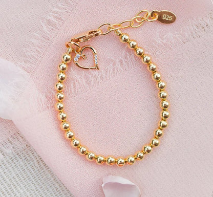 Close-up product shot of a gold beaded children’s bracelet arranged in a loop on pale pink fabric, featuring a lobster clasp, a dangling open‑heart charm with small crystals, an extension chain ending in an oval ‘925’ tag, and two soft pink rose petals.