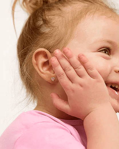Close-up of a smiling toddler in a pink shirt, holding her hands to her cheeks, wearing a small silver screw-back heart-shaped stud earring with a pink stone, against a pure white background