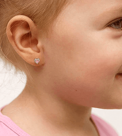 Close-up of a toddler’s ear wearing a small silver screw-back heart-shaped stud earring with a pink centre, set against a pure white background.