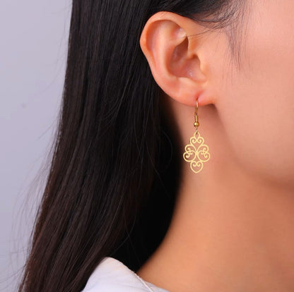 gold coloured surgical steel drop earring with an intricate pattern pictured on a young woman with dark hair on a light coloured background