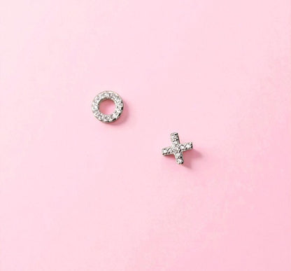 Two silver earrings, one circular and one with a 'X' design, on a pink background.