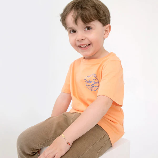 Child wearing an orange tee with Porto Surf graphic, showcasing a joyful smile and a cute identity bracelet.