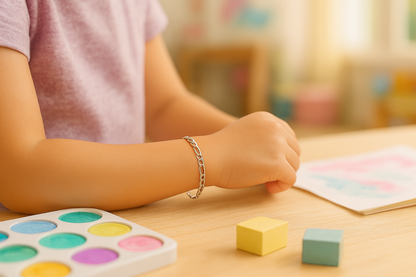 Close-up of a child’s forearm and hand resting on a light wood table, wearing a sterling silver Figaro chain bracelet; a watercolour palette and pastel-coloured wooden blocks sit nearby, with a softly blurred playroom in the background