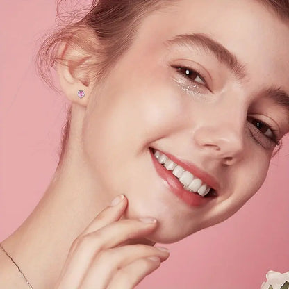 Close-up of a smiling young teen girl against a pink background, showcasing a small pink heart-shaped stud earring in her ear and wearing subtle sparkly eye makeup.