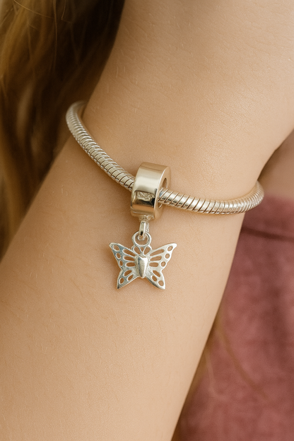 Close-up image of a young girl's wrist wearing a sterling silver snake chain bracelet with a dangling butterfly charm. The charm features delicate cut-out wing details and hangs naturally, with a soft-focus background showing the girl's light brown hair and a dusty rose top.
