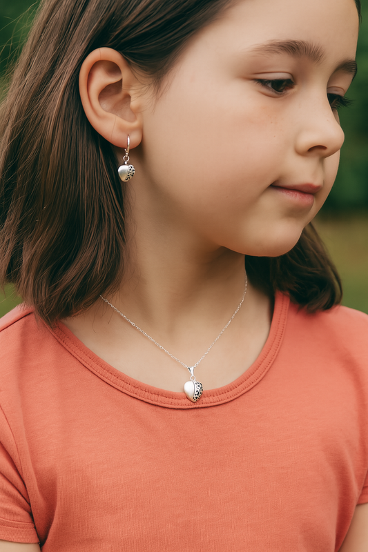 Close-up portrait of a young girl wearing a silver heart locket necklace with a half-polished, half-filigree design. She is also wearing matching earrings and a coral-coloured top, with a blurred green outdoor background.