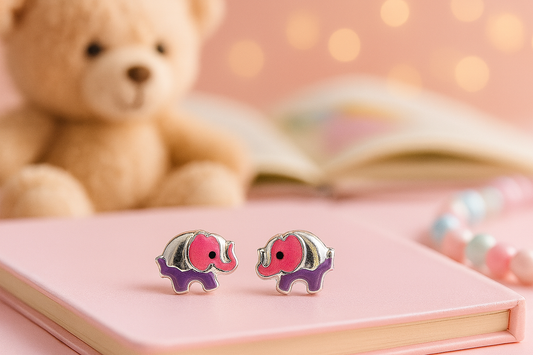 Elephant-shaped earrings on a pink surface with a teddy bear and book in the background