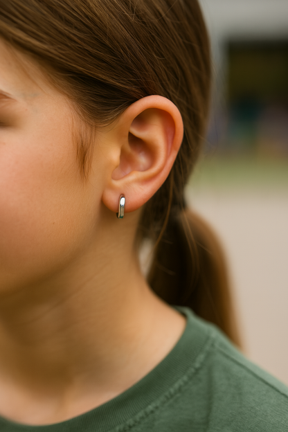 Close-up of an 8–12-year-old girl’s right ear wearing a small polished silver-tone hinged hoop earring, with her hair gently pulled back and a softly blurred outdoor background.