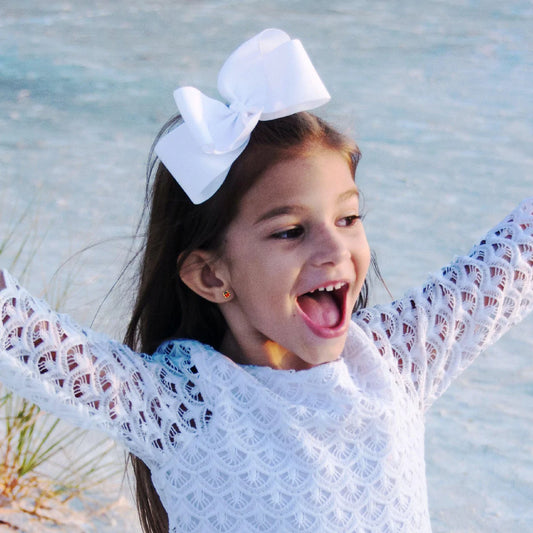 Cheerful girl wearing ladybug earrings with a big white bow excitedly celebrating at the beach, wearing a cute white lace top.