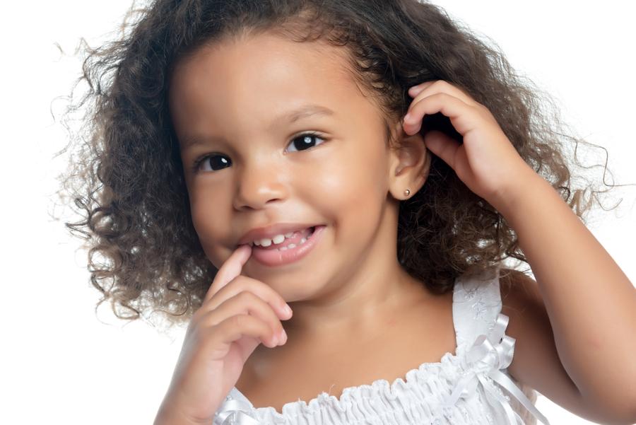 Cheerful young girl wearing gold ball stud earrings, smiling and holding her finger to her mouth.