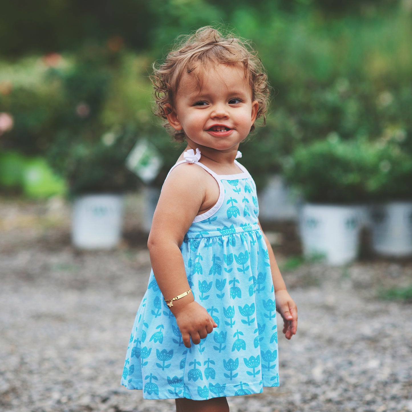 Smiling toddler wearing a blue dress and ID bracelet in a garden setting.