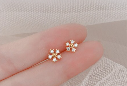 A pair of gold-plated children's earrings with snowflake flower designs and clear cubic zirconia, displayed on a person's fingertip.