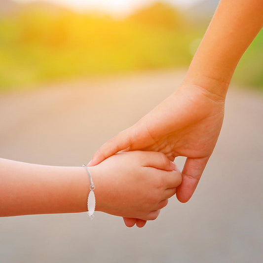 Close-up of an adult and child holding hands outdoors at sunset; the child wears a small sterling-silver ID bracelet with a scalloped oval name plate
