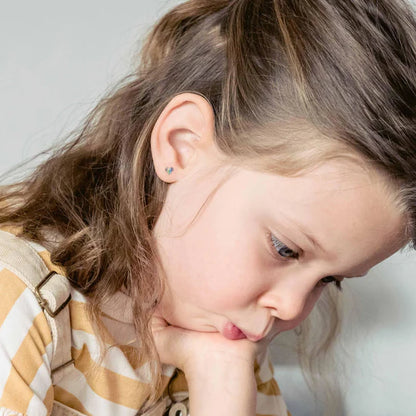 Close-up of a young girl’s left ear wearing a small sterling silver heart stud earring with rainbow-coloured vertical stripes.